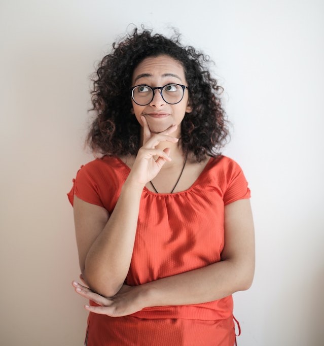 Girl with an orange shirt and black glasses standing against a white wall